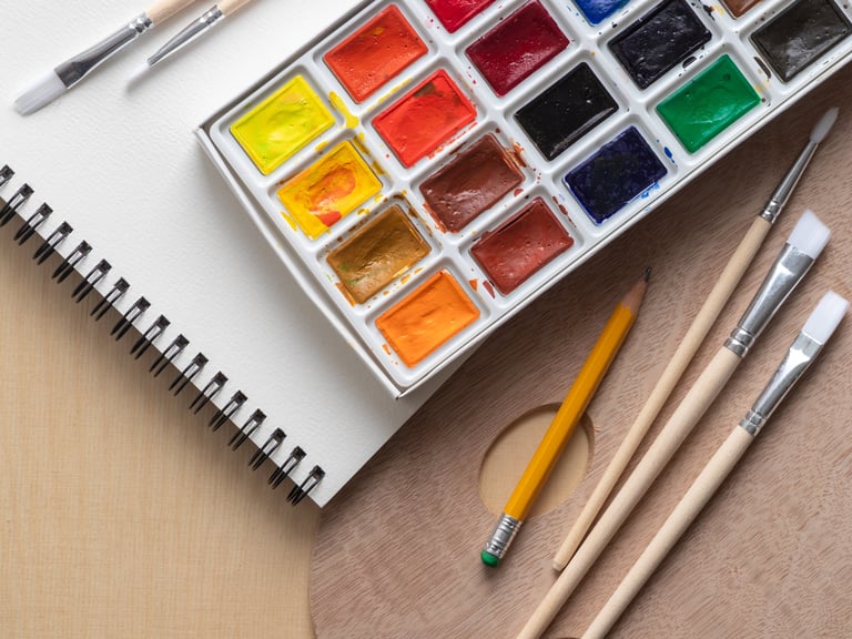 Watercolor art supplies on a light wooden surface, including paints, brushes, pencil, and spiral-bound sketchbook. Creative workspace for painting and sketching