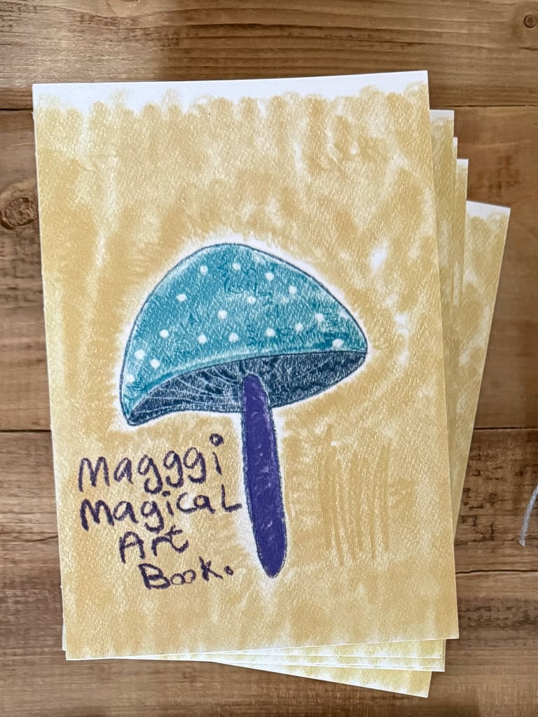 Hand-drawn illustration of a blue mushroom with purple stem on a cream-colored booklet cover titled Maggys Magical Art Book