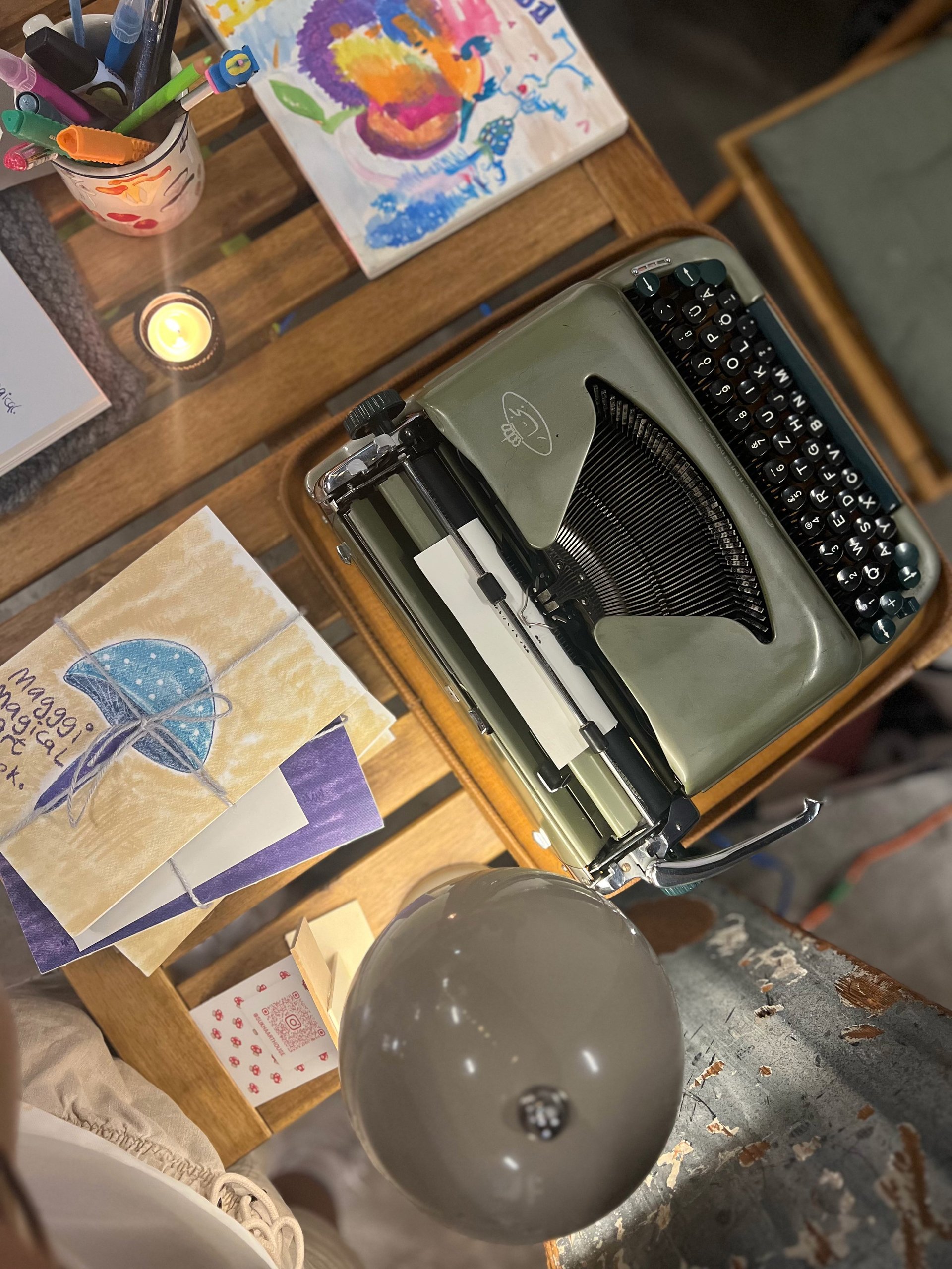 Vintage typewriter on wooden desk with art supplies, books, candle, and colorful artwork displayed above